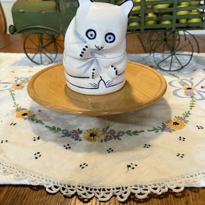 May include: A white ceramic panda-shaped container with blue accents, including eyes, ears, and decorative lines. The panda sits on a wooden plate atop a floral embroidered doily. A green metal toy truck is in the background.