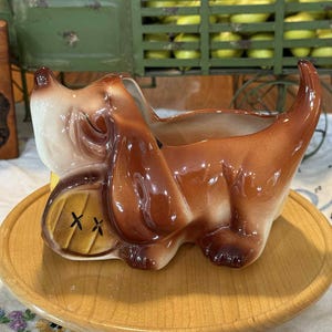 May include: A ceramic dog planter in shades of brown and cream, with a yellow barrel detail. The planter is sitting on a round wooden tray. The dog has floppy ears and a curved tail.