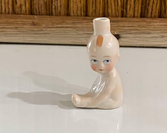 May include: A small porcelain figurine of a baby sitting with its legs crossed. The figurine is painted in a light pink color with blue eyes and a peach-colored blush on its cheeks.