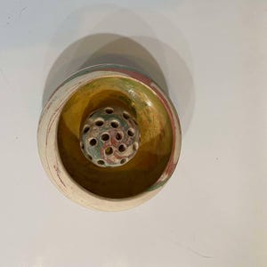 May include: A small, white ceramic bowl with a yellow and green glaze. The bowl has a round, open center with a smaller, round, green and brown ceramic ball with holes in it.