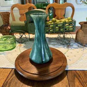 May include: A tall, green ceramic vase with a flared top sits on a round, wooden base. The vase has a glossy finish with darker green streaks. The wooden base is a rich brown color. The background includes a decorative cart and other vases.