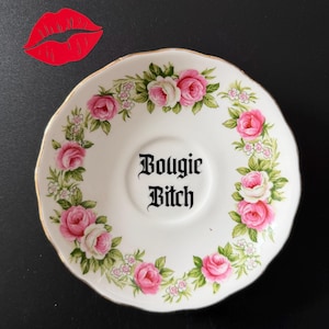 May include: A white ceramic saucer with a gold rim, decorated with pink roses and green foliage. The centre has the text "Bougie Bitch" in a gothic font. A red paper lip cutout is in the upper left corner.