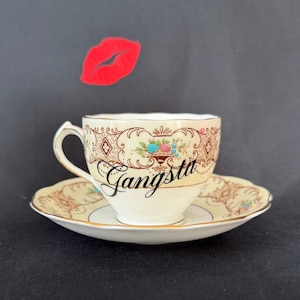 May include: A cream-coloured porcelain teacup and saucer set featuring a gold filigree pattern, a small fruit illustration, and the word Gangsta written in black script. A red lip icon is positioned above the teacup against a dark background.