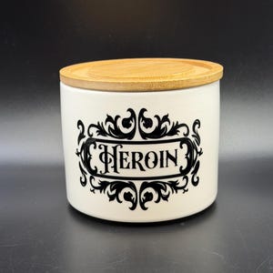 May include: A white ceramic jar with a bamboo lid. The jar features the word "HEROIN" in a black, ornate font, framed by decorative flourishes. The jar is cylindrical and sits on a dark surface.