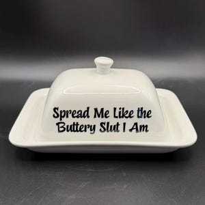 May include: White ceramic butter dish with a lid and a knob. The dish has the text "Spread Me Like the Buttery Slut I Am" in black lettering. The dish is rectangular with rounded corners.