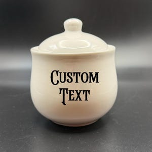 May include: A cream-coloured ceramic jar with a lid and a small knob on top. The jar has the words "CUSTOM TEXT" printed in black on the front. The jar is on a dark surface.