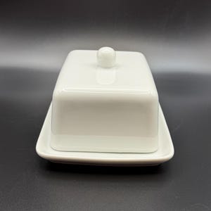 May include: A white rectangular butter dish with a lid and a small knob on top. The dish sits on a matching rectangular base. The design is simple and modern, with a glossy finish.