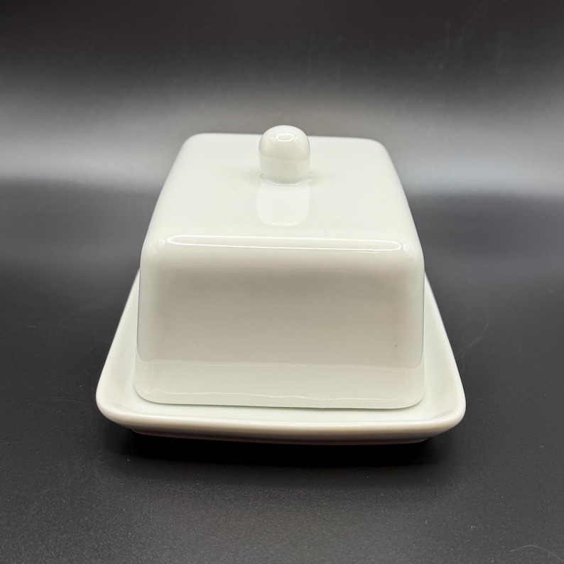 May include: A white rectangular butter dish with a lid and a small knob on top. The dish sits on a matching rectangular base. The design is simple and modern, with a glossy finish.