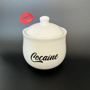 May include: A white ceramic jar with a lid, featuring the word "Cocaine" in black script. A red lip graphic is in the background. The jar has a rounded shape and a small knob on the lid.