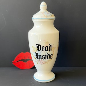 May include: A cream-coloured ceramic apothecary jar with a matching lid, featuring a floral pattern and the black text "Dead Inside" in a gothic font. The jar has a pedestal base and a pointed lid, both accented with thin light blue lines.