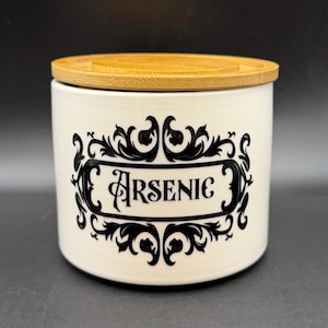 May include: An off-white ceramic jar with a bamboo lid. The jar has a black, ornate label that reads "Arsenic". The label is framed by decorative scrollwork. The jar is designed for storage.