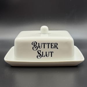 May include: A white ceramic butter dish with a lid and base. The front of the dish features the words "BUTTER SLUT" in black, stylized lettering. The lid has a small, round handle. The dish is rectangular with rounded corners.