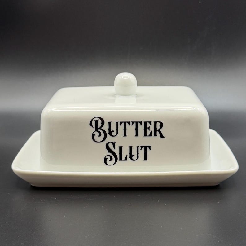 May include: A white ceramic butter dish with a lid and base. The front of the dish features the words "BUTTER SLUT" in black, stylized lettering. The lid has a small, round handle. The dish is rectangular with rounded corners.