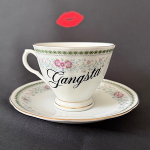 May include: A white porcelain teacup and saucer set featuring a delicate floral pattern in pink and green with gold trim. The word Gangsta is printed in black script on the front of the cup. A red lip icon is positioned above the teacup.