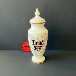 May include: A white ceramic apothecary jar with a matching lid, featuring a floral pattern in pastel pink and blue tones. The text "DEAD AF" is printed in bold black gothic lettering on the front. The jar has gold trim on the lid and base.