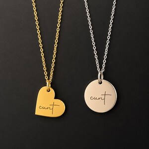 May include: Two necklaces with thin metal chains. One features a gold-colored heart-shaped pendant, and the other has a silver-colored circular pendant. Both pendants are engraved with the word "cunt" in a cursive, handwritten-style script font.