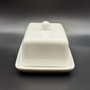 May include: A white ceramic butter dish with a lid and a small knob on top. The dish is rectangular and sits on a matching rectangular base. The design is simple and modern, suitable for kitchen use.