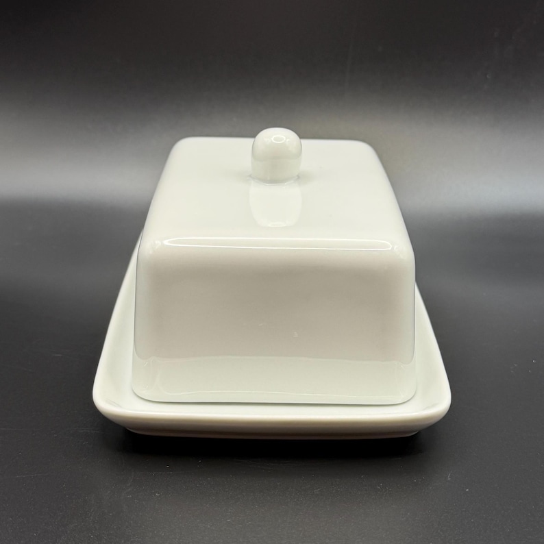 May include: A white ceramic butter dish with a lid and a small knob on top. The dish is rectangular and sits on a matching rectangular base. The design is simple and modern, suitable for kitchen use.