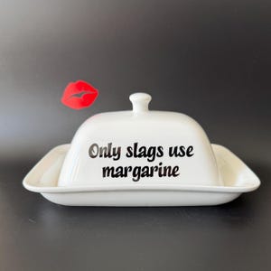 May include: White ceramic butter dish with a lid and a small handle. The dish has the text "Only slags use margarine" in black. A red lip sticker is in the background.