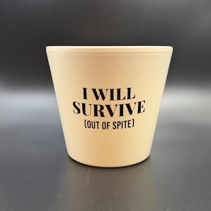 May include: A beige ceramic plant pot with the black text "I WILL SURVIVE (OUT OF SPITE)". The pot has a textured finish and a conical shape, wider at the top. The background is dark grey.
