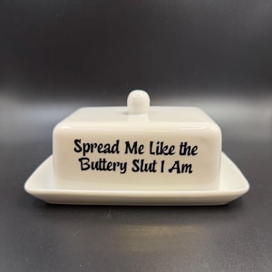 May include: A white ceramic butter dish with a lid and a small knob. The rectangular dish sits on a matching base. The text "Spread Me Like the Buttery Slut I Am" is printed in black on the front of the dish. A humorous kitchen item.