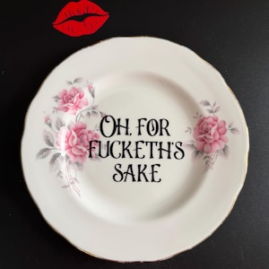 May include: A white ceramic plate with a scalloped edge and gold trim, featuring pink rose illustrations on the left and right sides. The centre of the plate displays the black text "OH, FOR FUCKETH'S SAKE" in a decorative, stylised serif font.