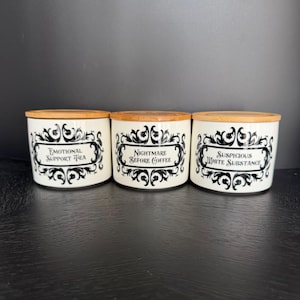 May include: Three white ceramic canisters with bamboo lids. Each canister has a black decorative label with text: "Emotional Support Tea", "Nightmare Before Coffee", and "Suspicious White Substance". The canisters are arranged on a dark surface.