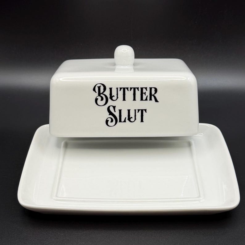 May include: A white ceramic butter dish with a lid and a base. The lid has a small, round handle and the words "BUTTER SLUT" in black script. The dish is rectangular and sits on a black surface.