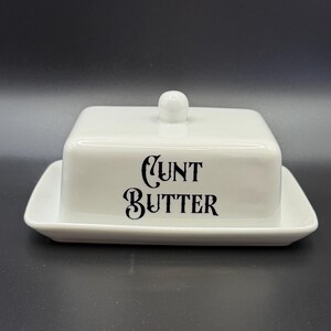 May include: A white ceramic butter dish with a lid and a small knob. The words "CUNT BUTTER" are printed in black, stylized lettering on the front of the dish. The dish sits on a rectangular base.
