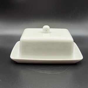 May include: A white ceramic butter dish with a square lid and a small, round knob. The dish sits on a rectangular base, all in a glossy white finish. The design is simple and elegant, suitable for kitchen or dining use.