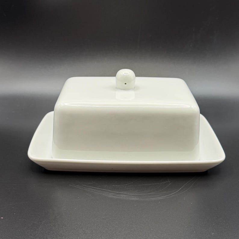 May include: A white ceramic butter dish with a square lid and a small, round knob. The dish sits on a rectangular base, all in a glossy white finish. The design is simple and elegant, suitable for kitchen or dining use.