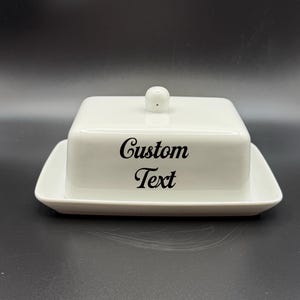 May include: A white ceramic butter dish with a lid and a base. The lid has a small, round knob. The words "Custom Text" are printed in black script on the front of the dish. The dish is rectangular with rounded corners.