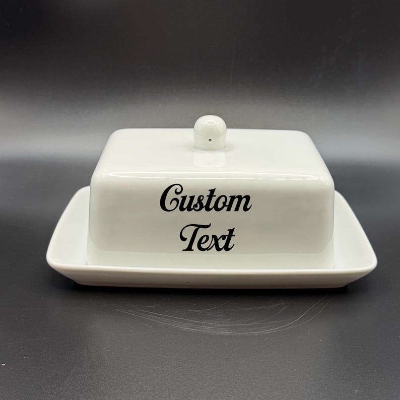 May include: A white ceramic butter dish with a lid and a base. The lid has a small, round knob. The words "Custom Text" are printed in black script on the front of the dish. The dish is rectangular with rounded corners.