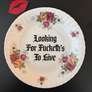 May include: A white ceramic plate with a scalloped edge, decorated with clusters of red and yellow roses. The center features black gothic-style text that reads, "Looking For Fucketh's To Give." A separate red lip-shaped sticker is visible above.