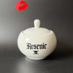 May include: A white ceramic lidded jar with a rounded body and a knob handle. The front features the word Arsenic printed in a black gothic-style font above a small black skull and crossbones icon. A red lip-shaped graphic floats above.