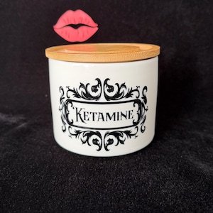 May include: White ceramic jar with a bamboo lid. The jar features a black ornate design with the word "KETAMINE" in the center. A pink lip graphic is above the jar.