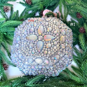 Pode incluir: Um ornamento esférico coberto com strass iridescentes de vários tamanhos e cristais maiores em forma de gota. A superfície apresenta um padrão de estrela. O texto Lucy e #1 está impresso em vermelho e branco na superfície do ornamento.