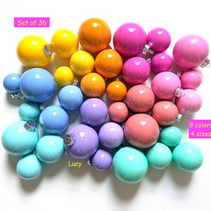 May include: A collection of 36 colorful Christmas ornaments in 9 colors and 4 sizes. The ornaments are arranged in a gradient of colors, including yellow, orange, pink, blue, and green. The text "Set of 36", "9 colors 4 sizes", and "Lucy" are visible.