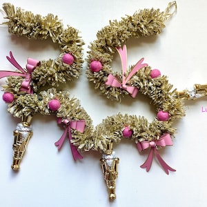 May include: Gold tinsel garland with pink bows and ornaments, and a gold nutcracker ornament. The garland is arranged in a circular shape. The word "Lucy" is written in pink.