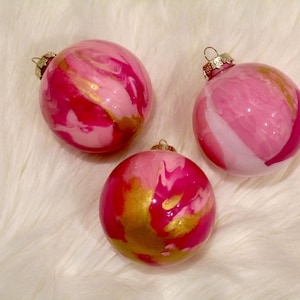May include: Three round Christmas baubles with a marbled design. The baubles are primarily pink with gold and white accents. Each bauble has a gold-coloured metal cap and a loop for hanging. The baubles are displayed on a white, fluffy surface.