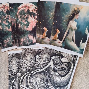 May include: A collection of art prints featuring fantasy themes. One print depicts a full moon over a forest and lake. Another shows a mermaid with a chihuahua. A third print is a black and white illustration of mermaids.