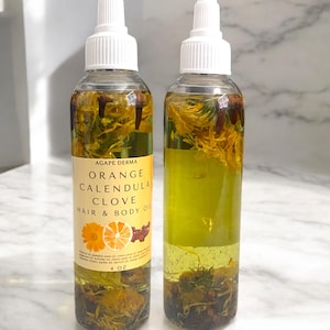 May include: Two clear glass bottles with white dropper tops filled with a yellow oil infused with orange slices, calendula flowers, and cloves. The bottles are labeled "Agape Derma Orange Calendula Clove Hair & Body Oil 4 oz."