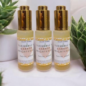 May include: Three facial serum bottles with gold-colored caps and labels that read "Turmeric & Carrot Brightening Facial Serum" from Agape Derma. The bottles are filled with a yellow liquid and are set against a white background with green plants.