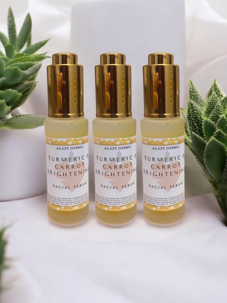 May include: Three facial serum bottles with gold-colored caps and labels that read "Turmeric & Carrot Brightening Facial Serum" from Agape Derma. The bottles are filled with a yellow liquid and are set against a white background with green plants.