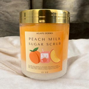 May include: A 12 ounce jar of peach milk sugar scrub with a gold lid. The label is orange and features a peach, a carton of milk, and a peach slice. The text on the label reads "Agape Derma Peach Milk Sugar Scrub".