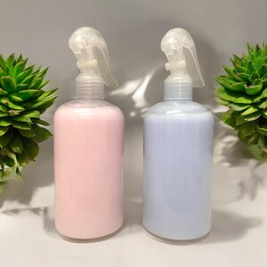 May include: Two clear plastic spray bottles, one filled with pink liquid and the other with blue liquid. Each bottle has a clear spray nozzle. Green succulent plants are on either side of the bottles.