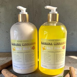 May include: Two bottles of Agape Derma Banana Cinnamon Oat Milk shampoo and conditioner, each 17 oz. The shampoo is yellow, the conditioner is white. Both have gold pumps and are labeled with text and banana and cinnamon illustrations. Cinnamon sticks are in the foreground.