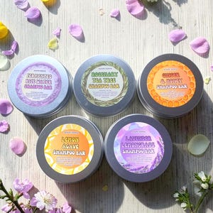 May include: Five round, silver shampoo bar tins with colorful labels and text. The labels read: Fermented Rice Water, Rosemary Tea Tree, Ginger & Honey, Lemon Agave, and Lavender Lemongrass. The image is decorated with flower petals and sprigs.