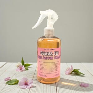 May include: A clear bottle of Agape Derma Guava Fig Rice Water Spray with a pink label. The bottle contains a light orange liquid and has a white spray nozzle. Pink flowers and petals are scattered around the bottle on a white wooden surface.