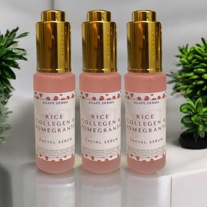 May include: Three pink facial serum bottles with gold-colored tops are displayed. Each bottle has a label with the text "RICE COLLAGEN & POMEGRANTE FACIAL SERUM". The bottles are arranged on a white surface with greenery in the background.
